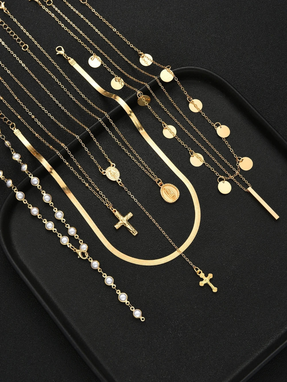 A set of gold and pearl religious necklaces and bracelets arranged on a black tray, featuring crosses, medallions, and bar charms.