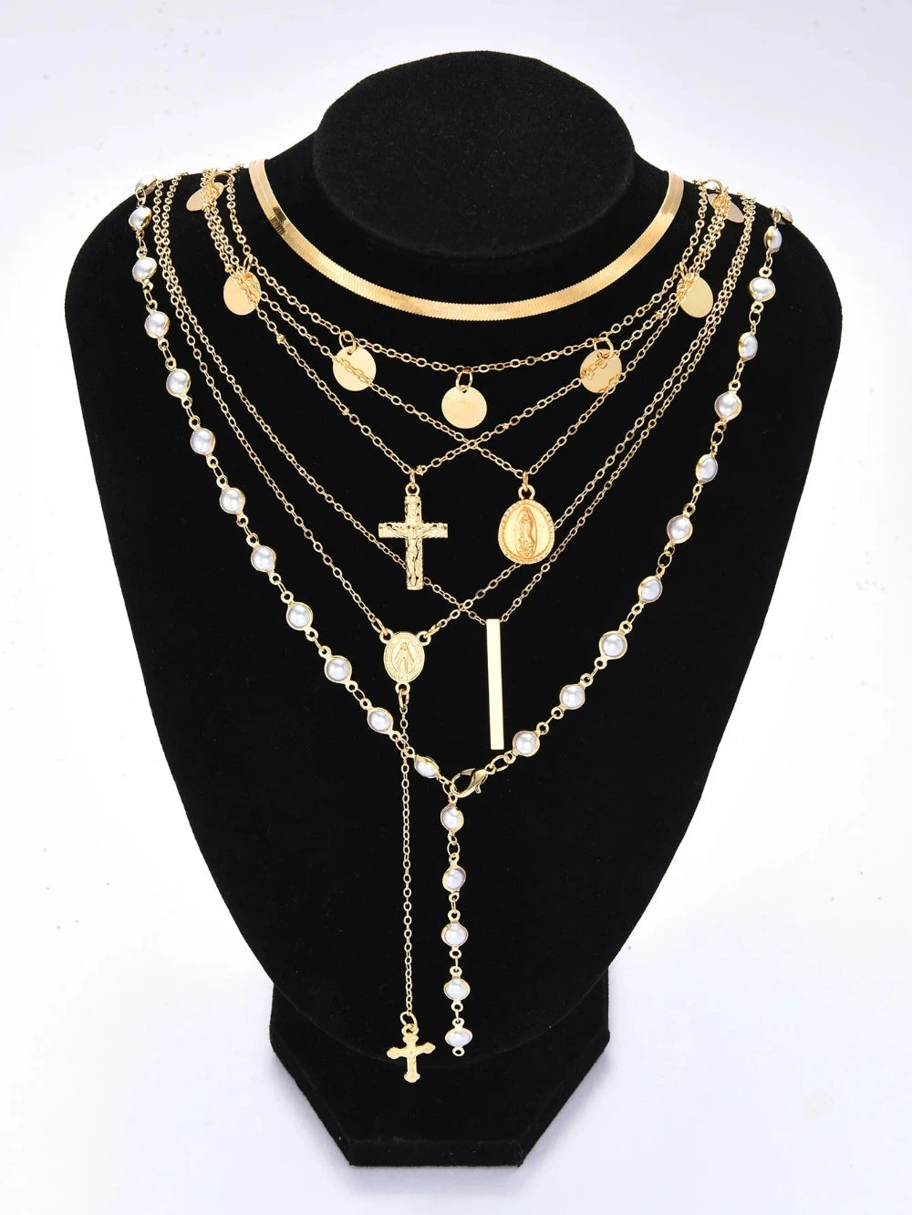 A multi-layered gold and pearl necklace displayed on a black velvet jewelry bust against a white background. The layered design features gold chains with round medallion pendants, a cross pendant, a vertical bar, and strands of white faux pearls.