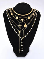 A multi-layered gold and pearl necklace displayed on a black velvet jewelry bust against a white background. The layered design features gold chains with round medallion pendants, a cross pendant, a vertical bar, and strands of white faux pearls.