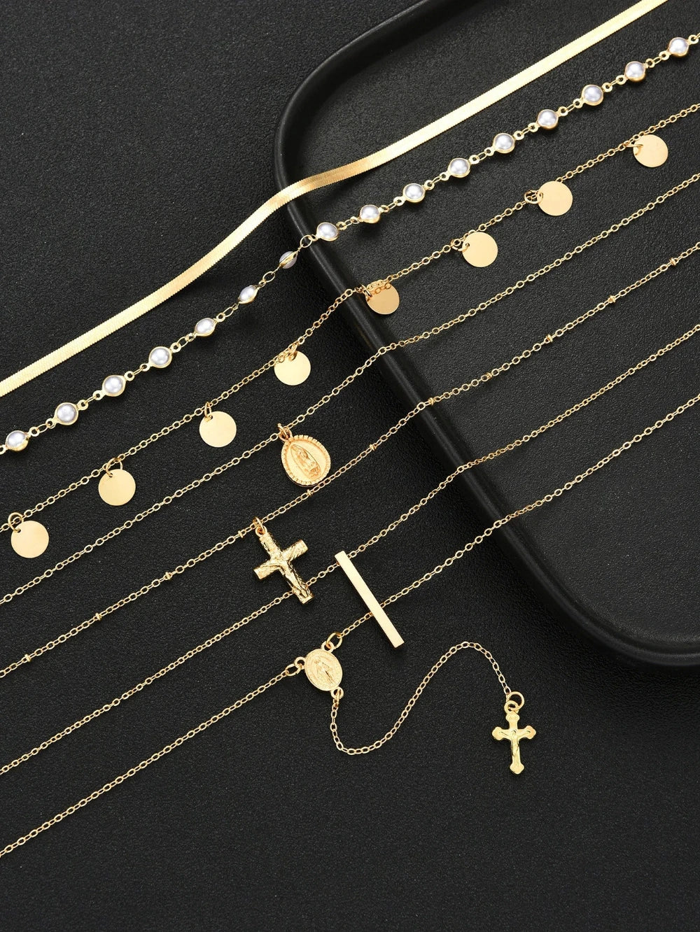 A set of gold-toned religious jewelry pieces, including necklaces with crosses, rosary-style chains, round and oval medallions, and pearl accents, arranged on a black surface next to a black jewelry box.