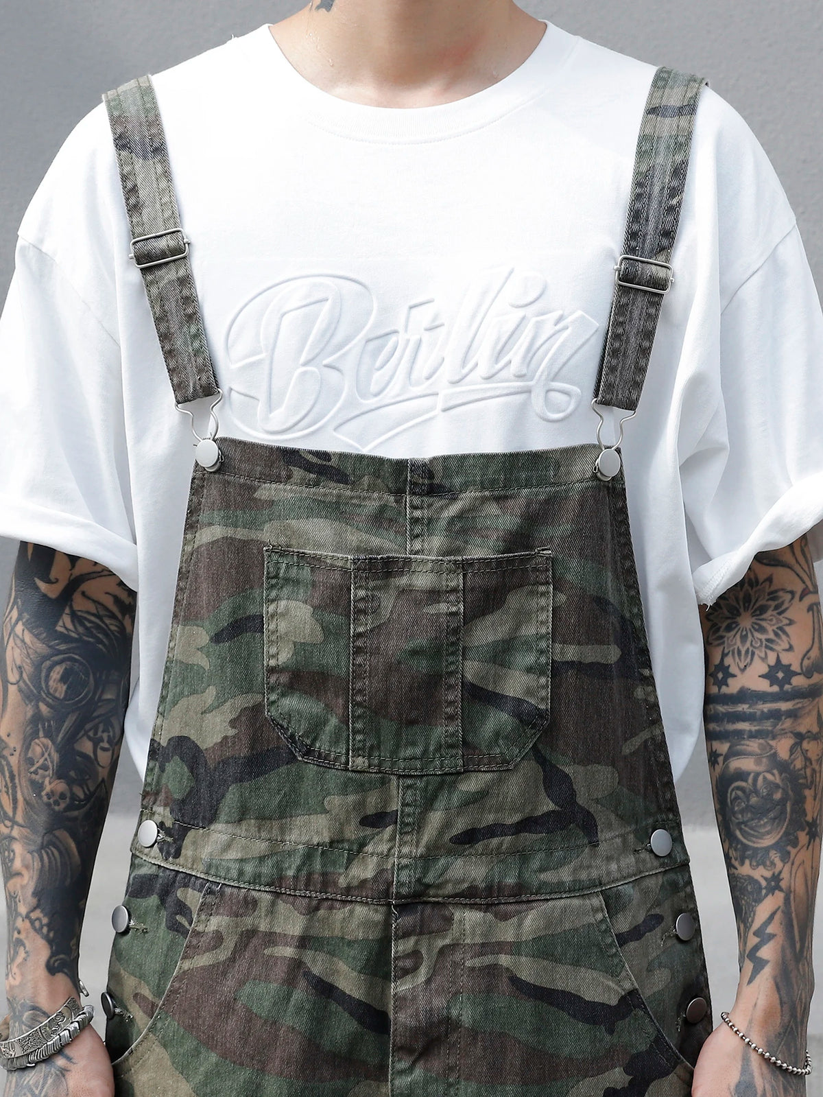 Person wearing a white t-shirt with 'Berlin' text and camouflage overalls.