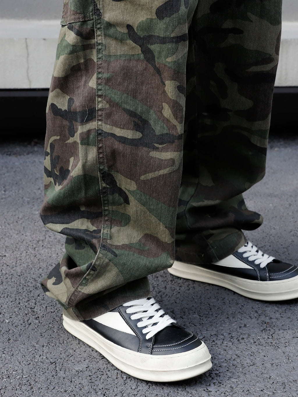 Person wearing camouflage pants and black and white sneakers on a concrete surface