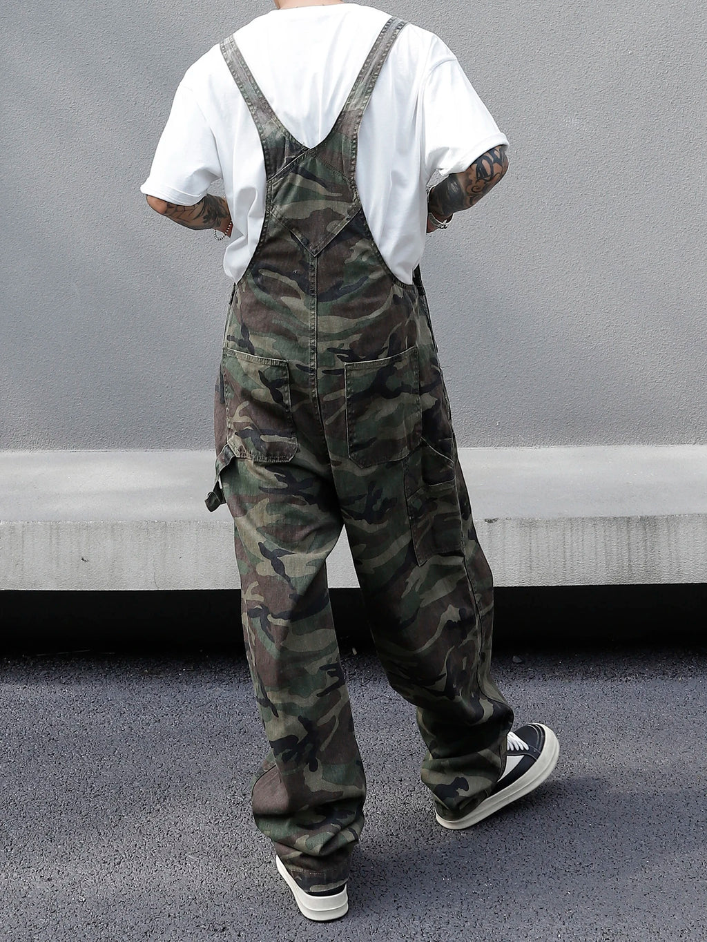 Person wearing camouflage overalls with a white shirt underneath, standing against a gray wall.
