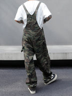 Person wearing camouflage overalls with a white shirt underneath, standing against a gray wall.