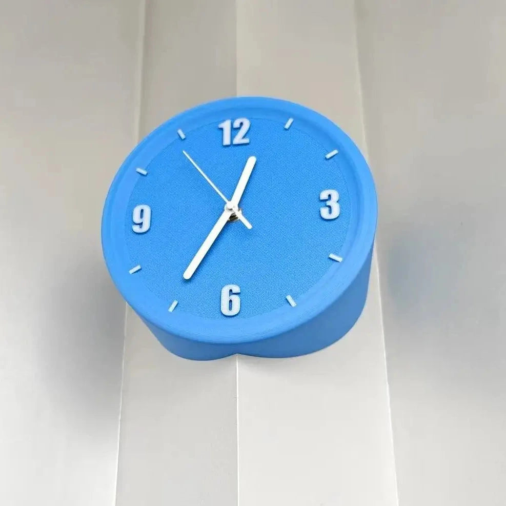 A blue heart-shaped wall clock with white hands and white Arabic numerals, mounted on a white paneled wall.