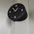 A modern black cylindrical wall clock with white numerals and hands, mounted on a light gray paneled wall.