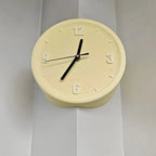 A round wall clock with a pale cream-colored textured body, black hands, and raised Arabic numerals, mounted on a white paneled wall.