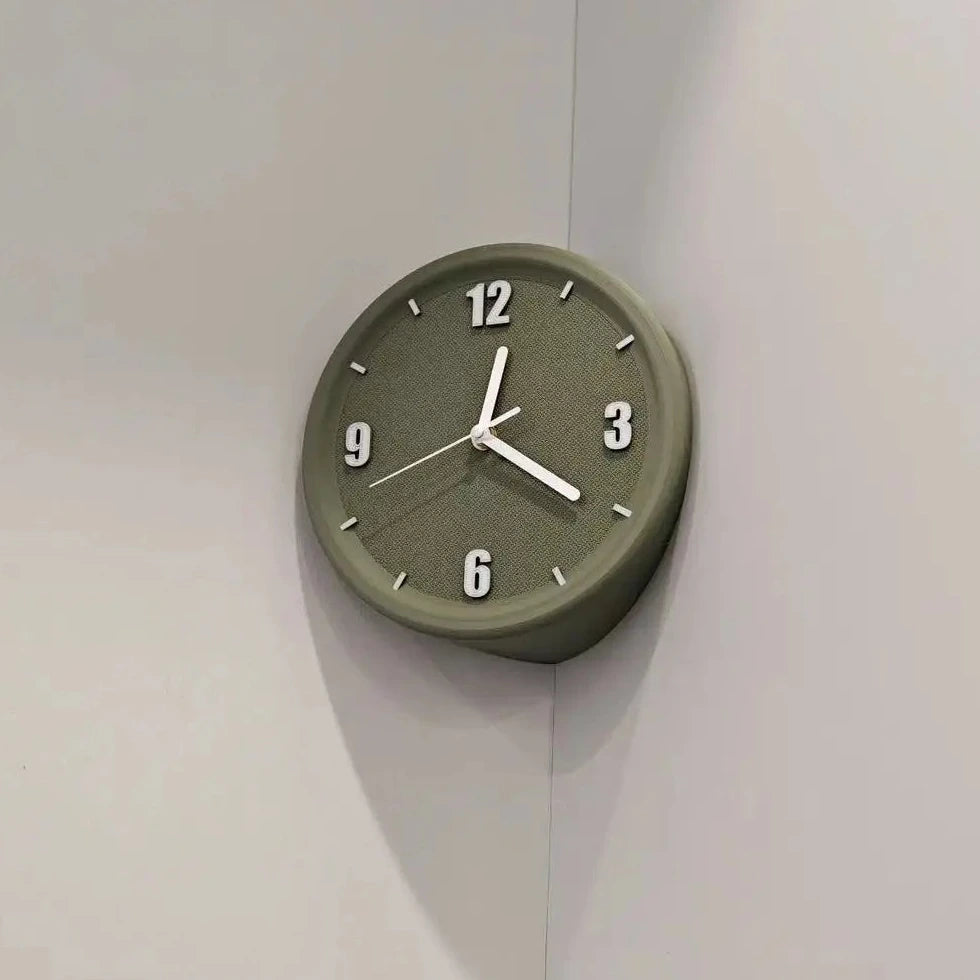 An olive green round wall clock with white hands and black Arabic numerals, mounted on a light gray corner wall.