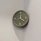 An olive green round wall clock with white hands and black Arabic numerals, mounted on a light gray corner wall.