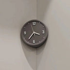 A minimalist round wall clock with a dark gray face and matching outer rim, featuring white Arabic numerals and hands, mounted on a light gray corner wall.