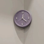A round wall clock with a muted lavender-purple face and white hands, mounted on a light beige corner wall.