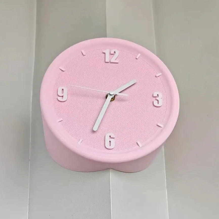 A pink heart-shaped wall clock with white hands and white Arabic numerals, mounted on a white paneled wall.