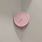A round pink wall clock with black Arabic numerals and white hands, mounted on a light gray corner wall.