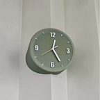 A round wall clock in a muted sage green color, mounted on a white paneled wall. The clock features white Arabic numerals and white hands.