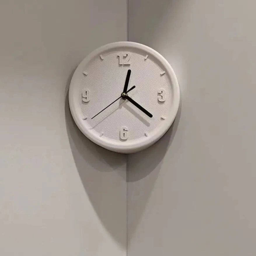 A minimalist white round wall clock with black hands and numerals, mounted on a light gray corner wall.