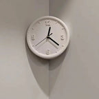 A minimalist white round wall clock with black hands and numerals, mounted on a light gray corner wall.