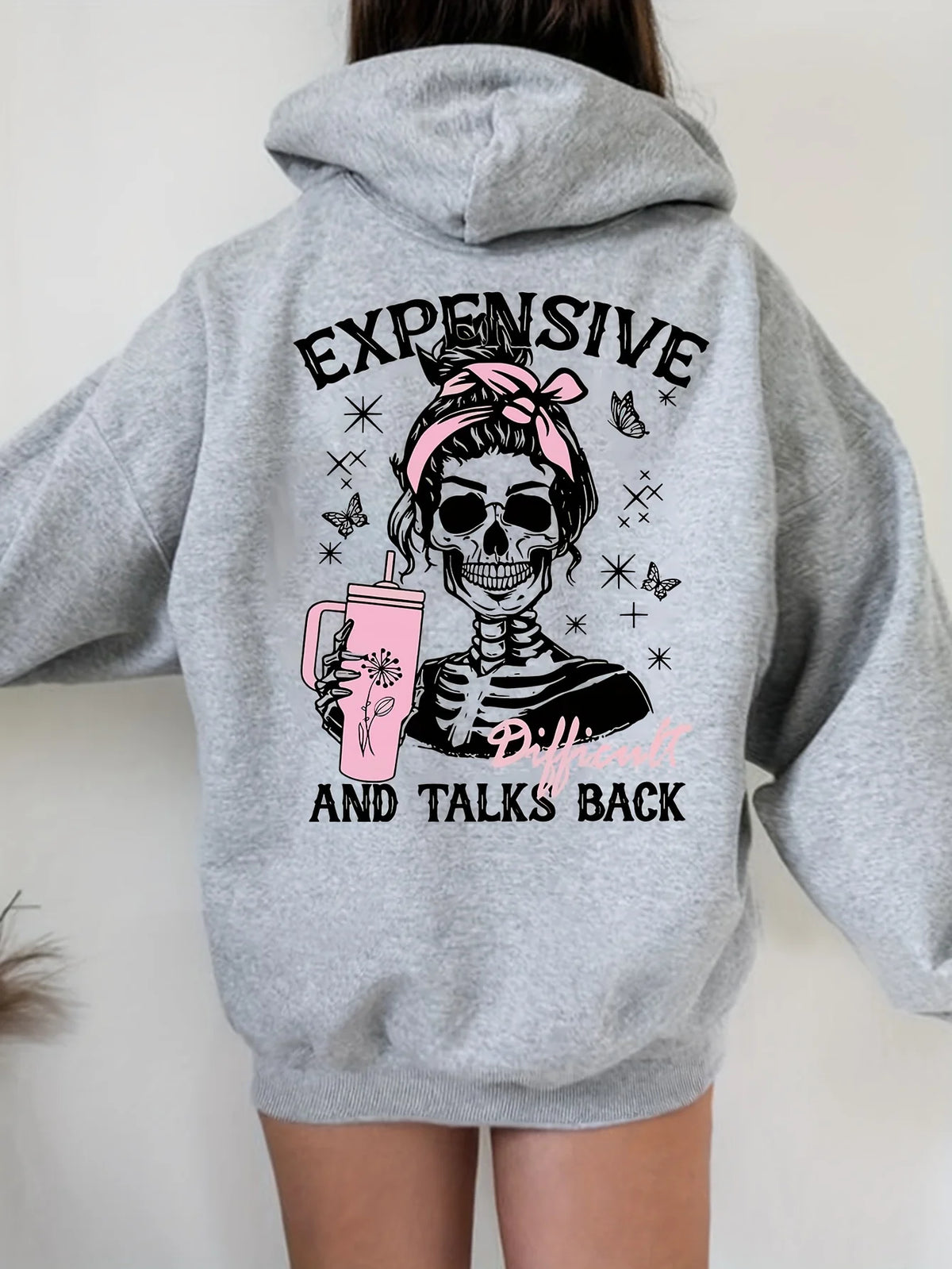 A person wearing a heather gray hoodie from the back, featuring a graphic print of a skeleton woman with a pink bow and a pink drink, with the text 'EXPENSIVE' and 'AND TALKS BACK'.