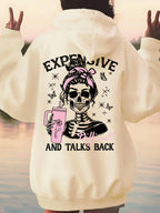 A person shown from the back wearing an oversized cream hoodie with a large graphic print on the back. The design features a cartoon skeleton with a pink hair bow, holding a pink drink, surrounded by butterflies and stars, with the text 'EXPENSIVE' at the top and 'AND TALKS BACK' at the bottom.