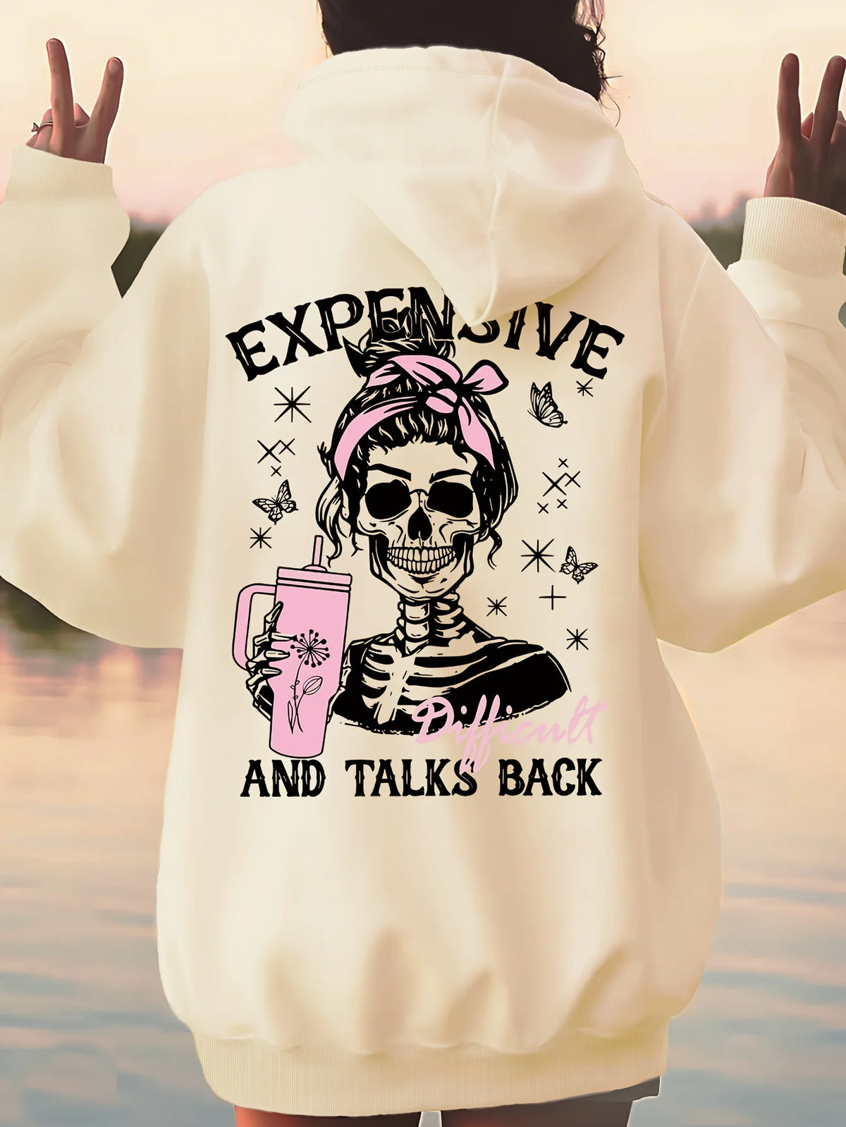 A person shown from the back wearing an oversized cream hoodie with a large graphic print on the back. The design features a cartoon skeleton with a pink hair bow, holding a pink drink, surrounded by butterflies and stars, with the text 'EXPENSIVE' at the top and 'AND TALKS BACK' at the bottom.