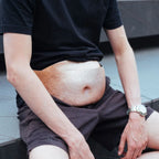 A man wearing a black t-shirt and dark brown shorts, sitting on outdoor steps with his shirt pulled up to expose his stomach. The stomach area is digitally blurred/censored.