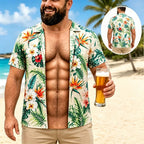 A man on a beach wearing a short-sleeve open button-up Hawaiian shirt with a tropical floral print. The shirt has green palm leaves and orange, red, and white flowers on a light beige background.