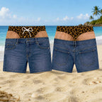 A pair of denim shorts shown from the front and back on a sandy beach with ocean and palm trees in the background. The shorts have a leopard-print waistband with a white bow on the front view and a tan panel on the back view.