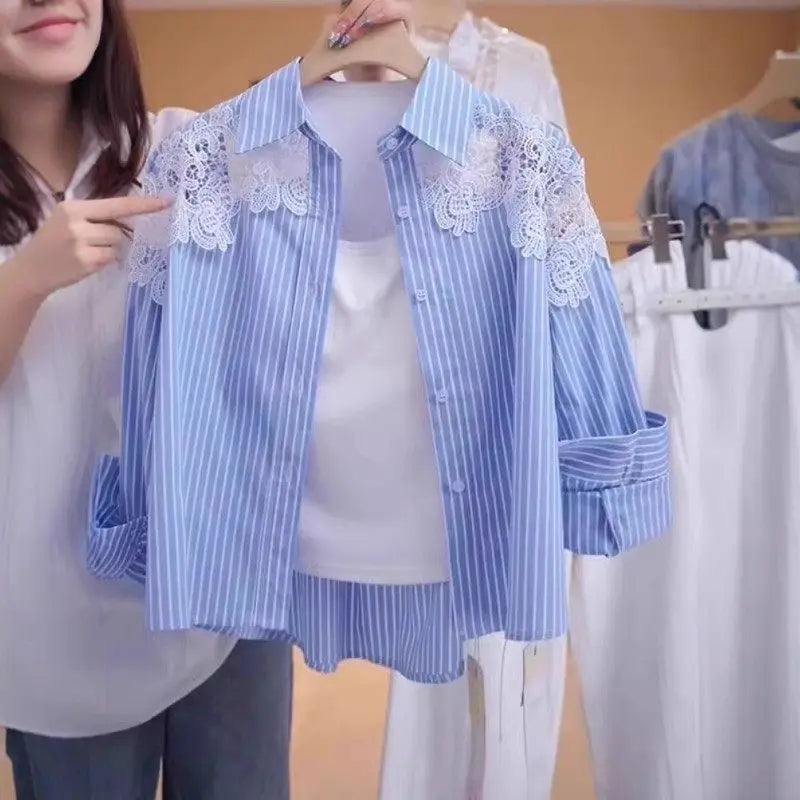 A woman holds up a light blue and white vertically striped button-up shirt with white floral lace appliques on the shoulders, layered over a white tank top. The background shows a clothing rack with other garments.