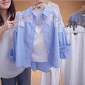 A woman holds up a light blue and white vertically striped button-up shirt with white floral lace appliques on the shoulders, layered over a white tank top. The background shows a clothing rack with other garments.