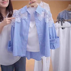 A woman holds up a light blue and white vertically striped button-up shirt with white floral lace appliques on the shoulders, layered over a white tank top. The background shows a clothing rack with other garments.