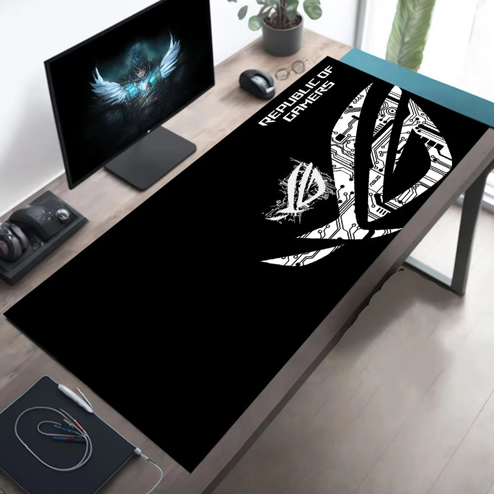 A black desk mat with a white Republic of Shadows logo and text, laid out on a wooden desk in a modern home office setup alongside a computer monitor, mouse, and headset.