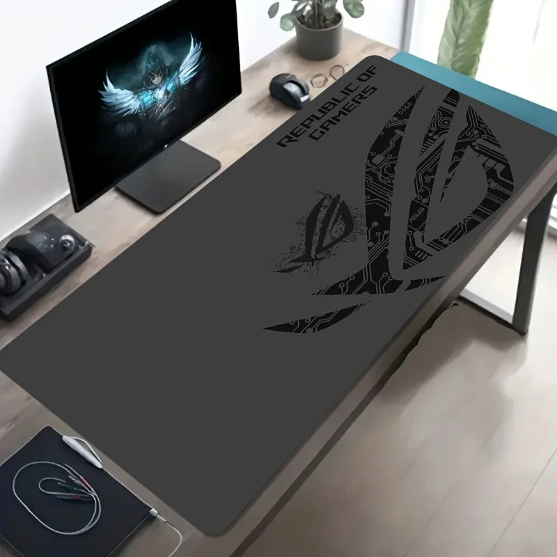 A large dark gray gaming desk mat with a black Republic of Gamers logo and stylized emblem, shown on a desk alongside a monitor, mouse, and headset in a home office setting.