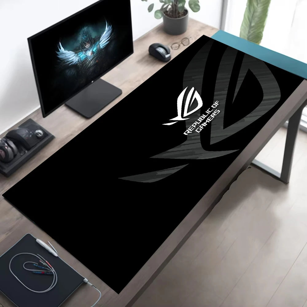 A large black gaming desk mat with a white Republic of Shadows logo, shown on a wooden desk in a modern home office setup alongside a computer monitor, mouse, and headset.