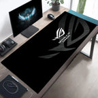 A large black gaming desk mat with a white Republic of Shadows logo, shown on a wooden desk in a modern home office setup alongside a computer monitor, mouse, and headset.