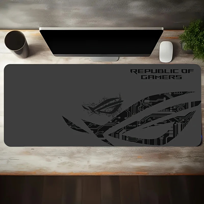 A large dark gray gaming desk mat with a black Republic of Gamers logo and circuit-style graphic, shown on a wooden desk with a monitor, mouse, and cup nearby.