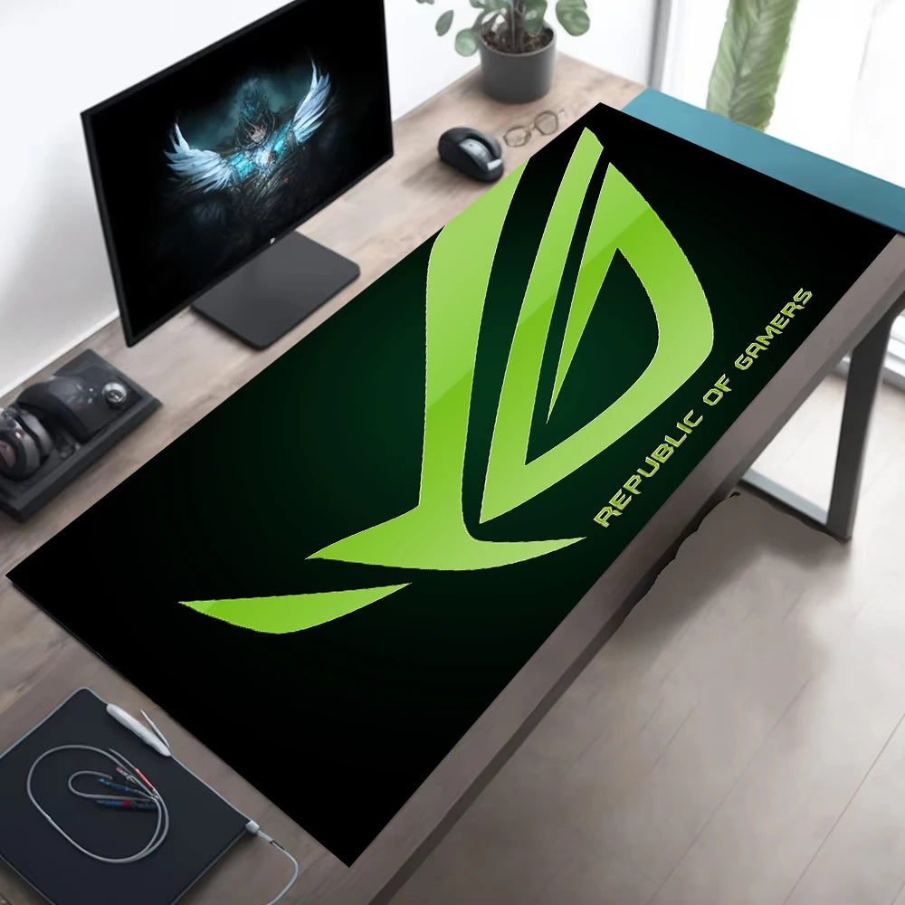 A large black desk mat with a bright green Republic of Gamers logo and text, shown on a wooden desk in a modern home office setup alongside a computer monitor, mouse, and other desk accessories.