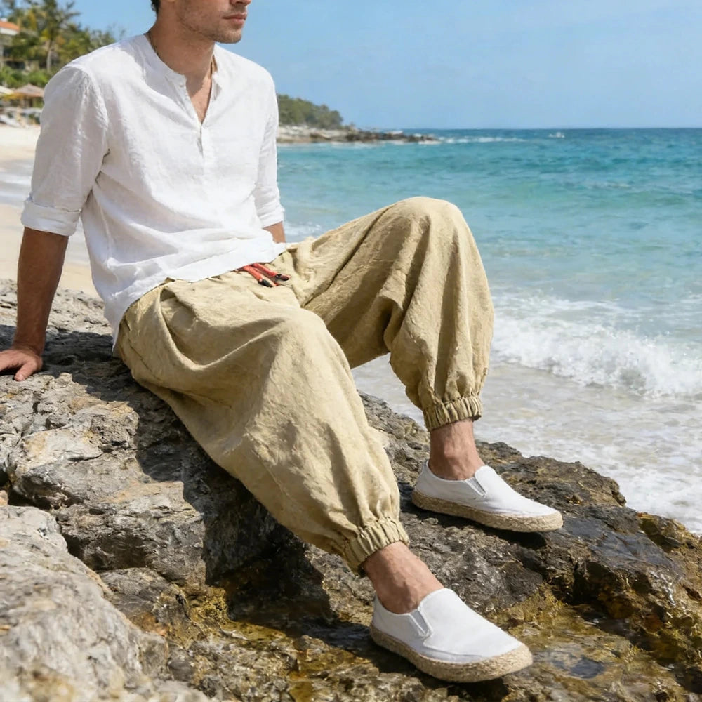 Man sitting on rocks by the ocean wearing a white shirt, beige pants, and white shoes.