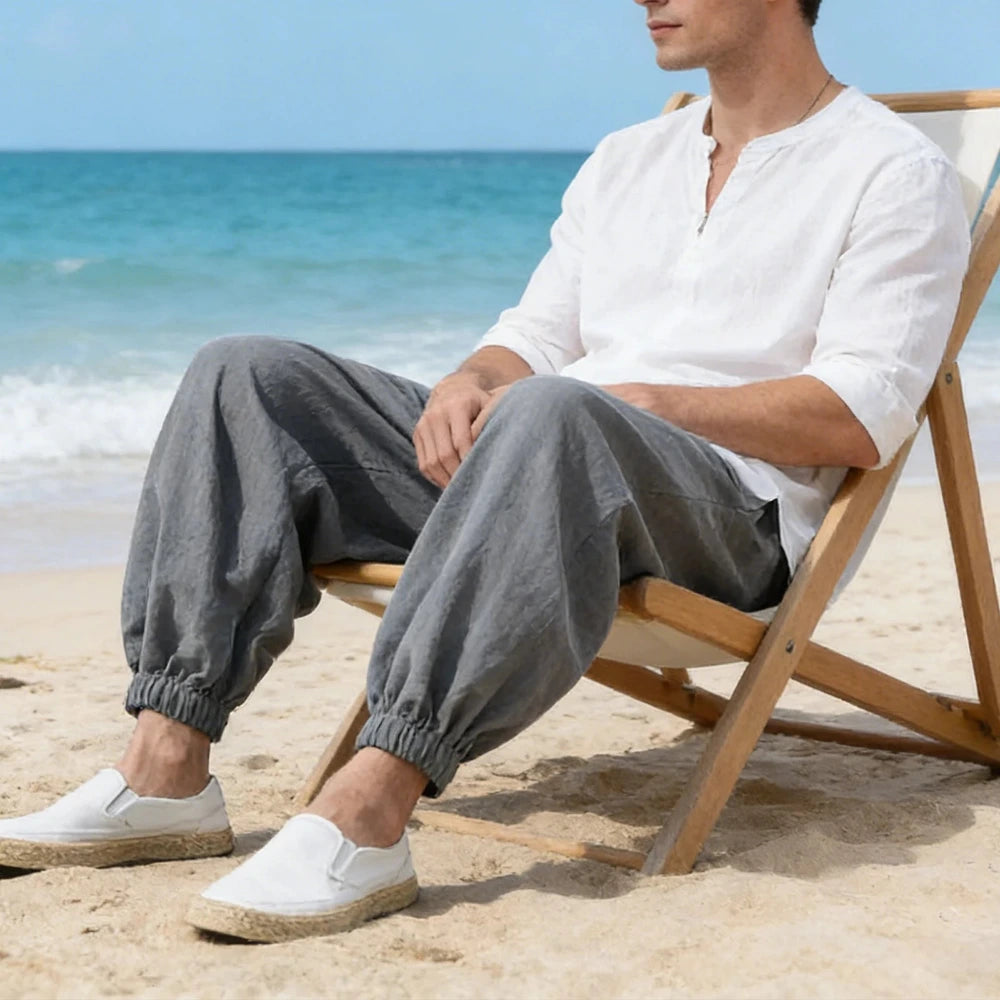 Man sitting on a beach chair by the ocean wearing a white shirt and gray pants.