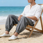 Man sitting on a beach chair by the ocean wearing a white shirt and gray pants.