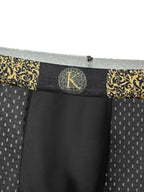 Close-up of men's boxer briefs showing the waistband with a black and gold damask pattern, a central circular monogram logo with a glittery gold 'K', and a black front pouch.