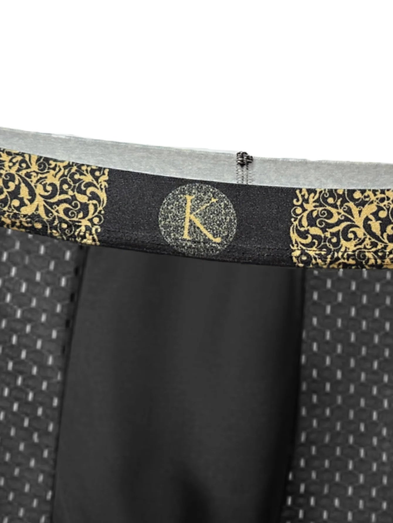 Close-up of men's boxer briefs showing the waistband with a black and gold damask pattern, a central circular monogram logo with a glittery gold 'K', and a black front pouch.