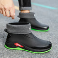Black rain boot with gray cuff and green sole on a gray pavement background