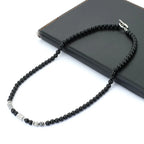 A beaded necklace made of small black round beads with a few silver and gray spacer beads, displayed on a black rectangular surface against a white background.