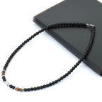 A beaded necklace with predominantly black round beads, accented by a silver-toned cylindrical bead and two small brown tiger's eye beads, displayed on a black leather journal against a white background.