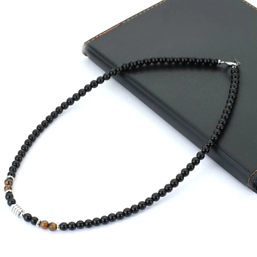 A beaded necklace with predominantly black round beads, accented by a silver-toned cylindrical bead and two small brown tiger's eye beads, displayed on a black leather journal against a white background.