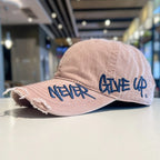 Pink cap with 'Never Give Up' text on a blurred indoor background