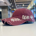 Maroon cap with 'Never Give Up' text on a blurred indoor background