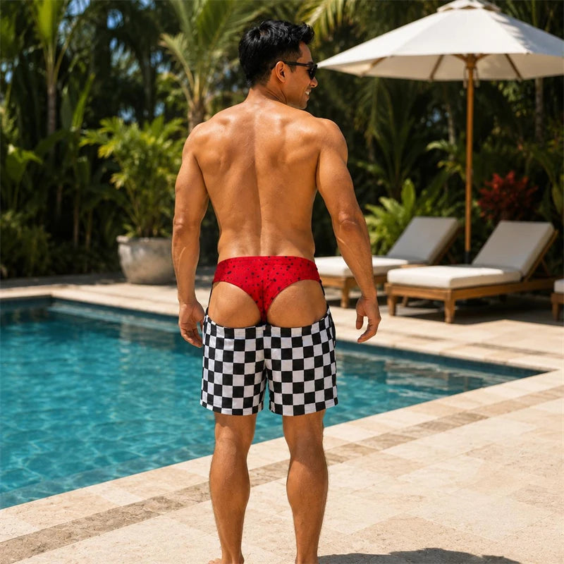 A man standing by a swimming pool wearing red swim briefs with a small black pattern and black-and-white checkered swim trunks, photographed outdoors with lounge chairs and palm trees in the background.