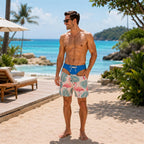 A man standing on a sandy beach wearing blue swim briefs with a pink flamingo and green palm leaf print, photographed outdoors with ocean, trees, and lounge chairs in the background.