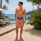 A man standing on a sandy beach wearing patterned swim trunks and a purple swim brief, with a tropical ocean and palm trees in the background.