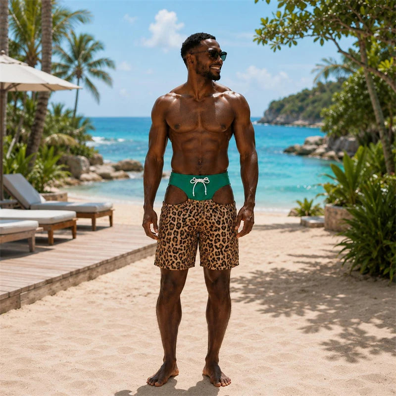 A muscular man stands on a sandy beach wearing leopard-print swim trunks with a bright green waistband and white drawstring, with turquoise ocean, palm trees, and lounge chairs in the background.